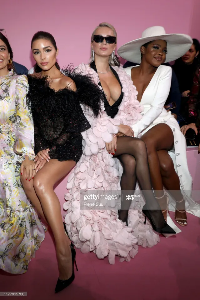 Ryan Destiny sitting with guests at a fashion show in a white outfit and heels.