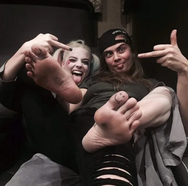 Cara Delevingne posing with a companion with bare feet raised.