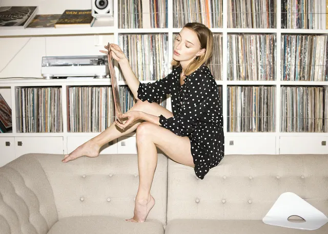 Phoebe Dynevor seated beside records in a black polka-dot dress.
