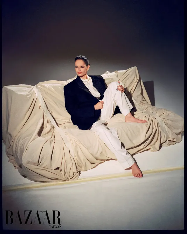 Eva Green seated on a draped sofa in a dark jacket.