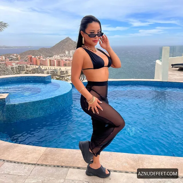 Cierra Ramirez in a black bikini top, sheer trousers and dark flats posing beside a rooftop pool.