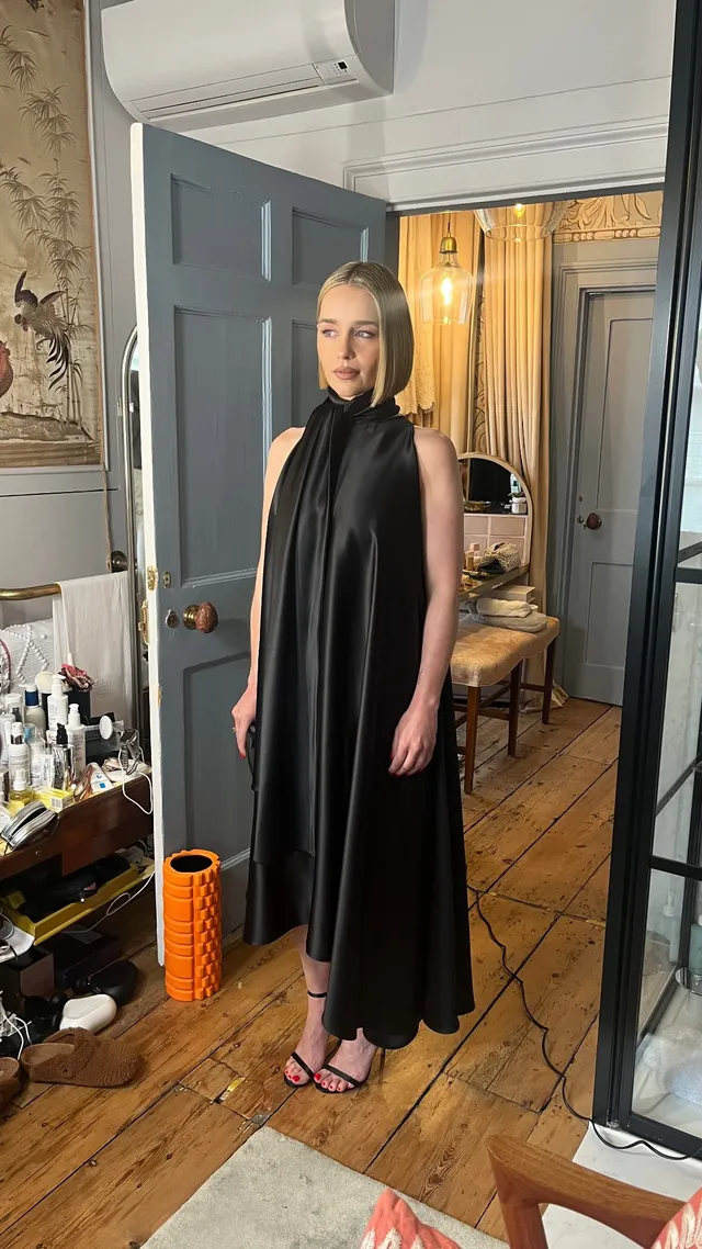 Emilia Clarke standing by mirrors in a black dress and heels.