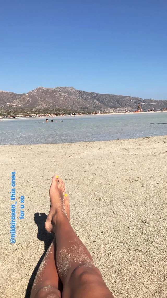 Ella Purnell relaxing on a sandy beach with one leg crossed.