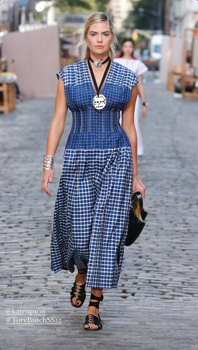 Kate Upton walking outside in a checked dress and black heels.