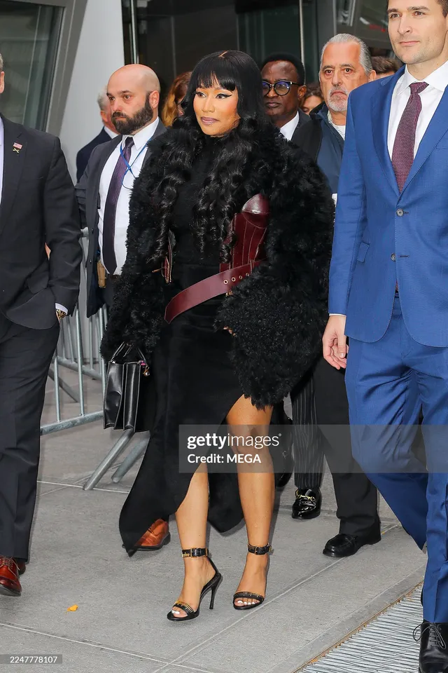 Nicki Minaj walking beside other people in a dark outfit and black heels.