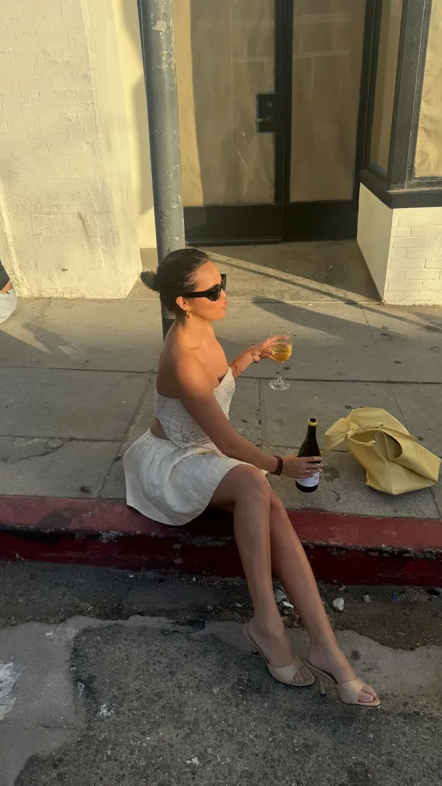 Bailee Madison sitting outdoors in a white dress and flat slides.