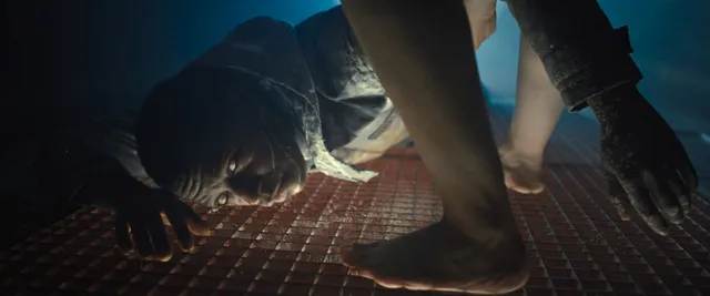 Cailee Spaeny standing barefoot on a grated floor in a film scene.