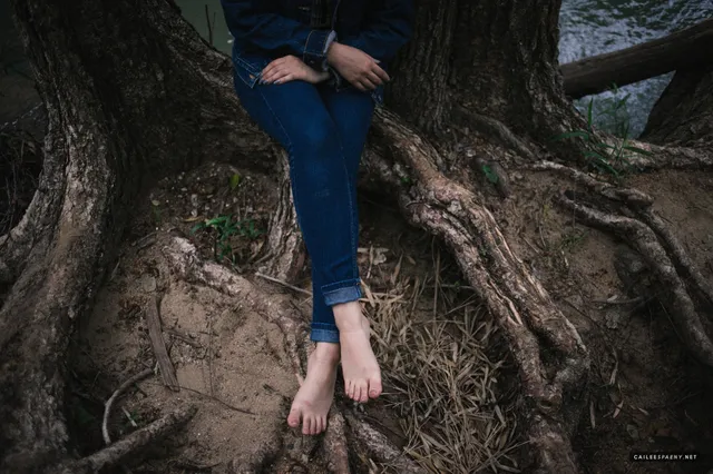 Cailee Spaeny sitting barefoot in blue jeans beside a tree.