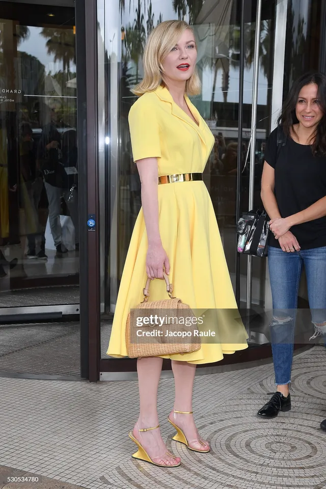 Kirsten Dunst wearing a yellow dress and wedge heels on a sidewalk.