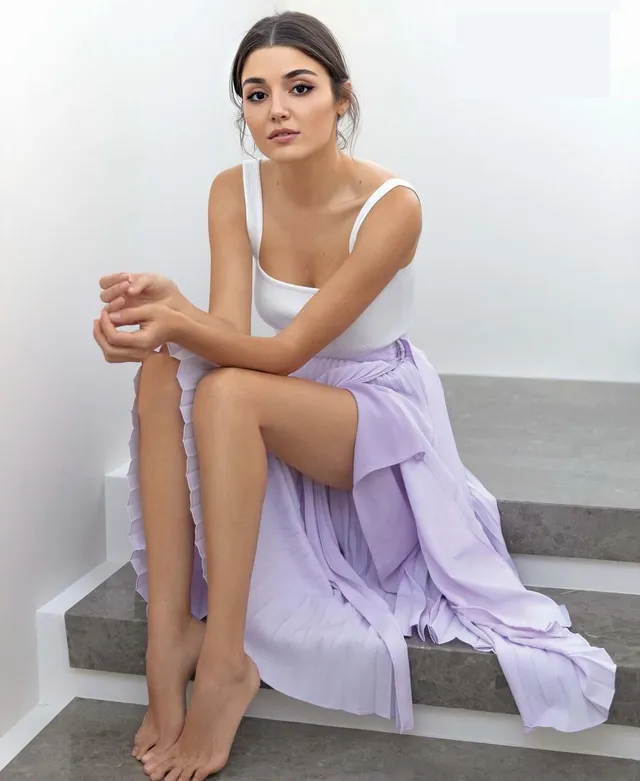 Hande Erçel sitting barefoot on steps in a white top and lavender skirt.