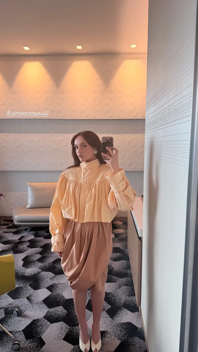 Olivia Cooke taking a mirror photograph in a loose blouse and skirt.