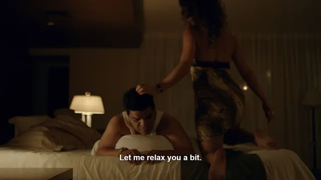 Adria Arjona standing beside a man on a bed in a film scene.