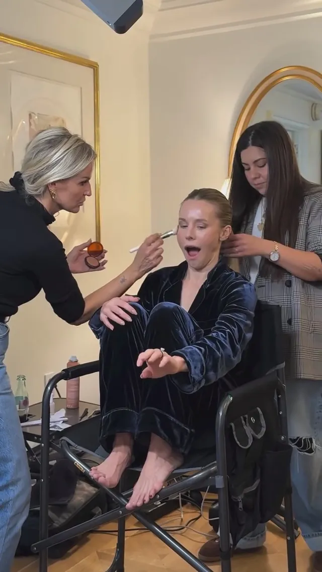Kristen Bell having makeup applied while seated barefoot in a chair.