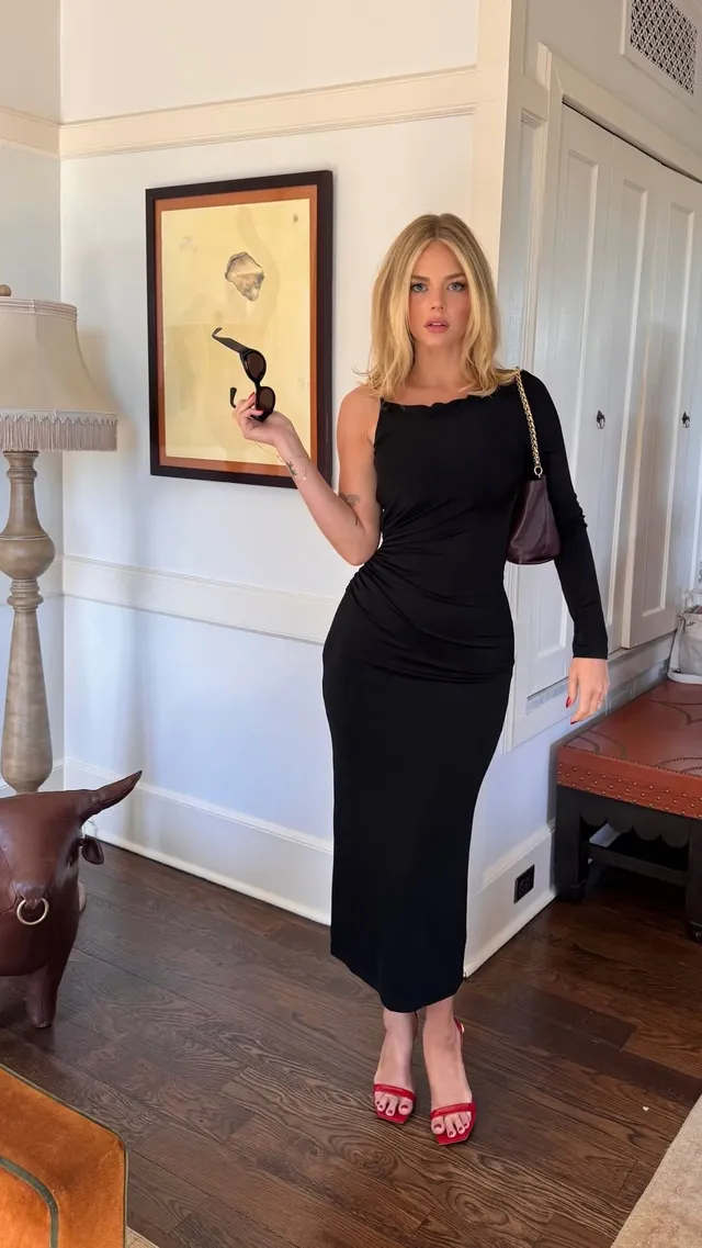 Samara Weaving posing indoors in a black dress and red heels.