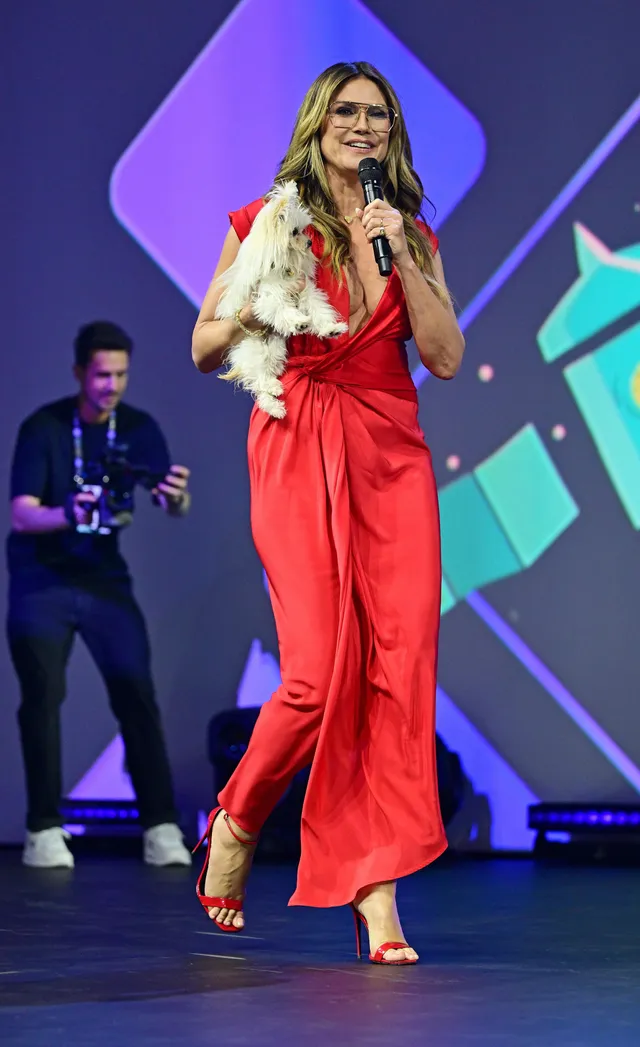 Heidi Klum standing on stage with a microphone in a red wide-leg jumpsuit and heels.