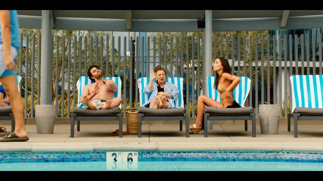 Kaley Cuoco sitting barefoot beside a swimming pool with other people.