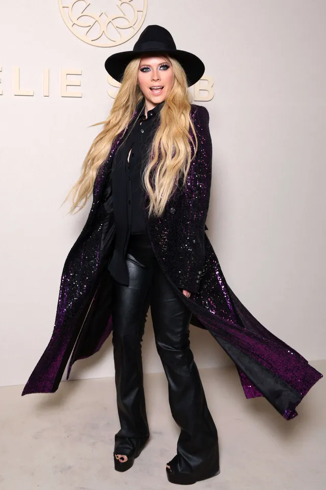 Avril Lavigne standing at an event in a long coat and platform heels.