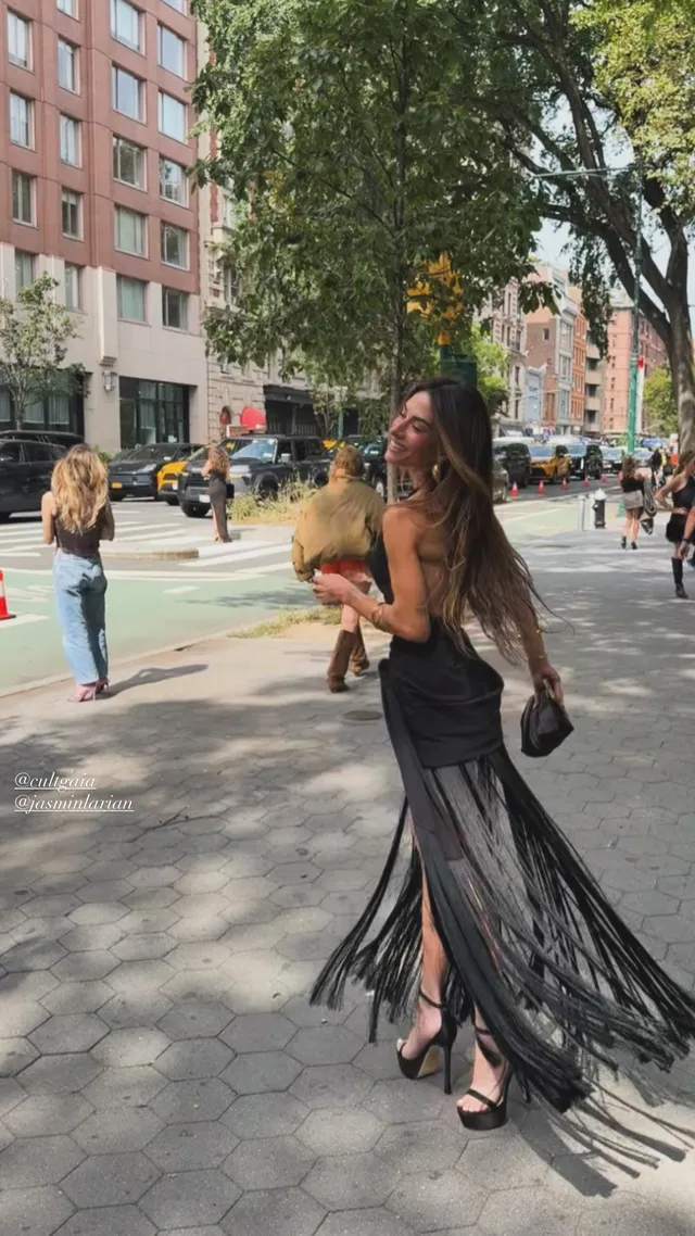 Sarah Shahi walking on a city pavement in a long dark outfit.