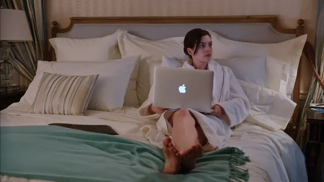 Anne Hathaway sitting on a bed with a laptop and bare feet visible.