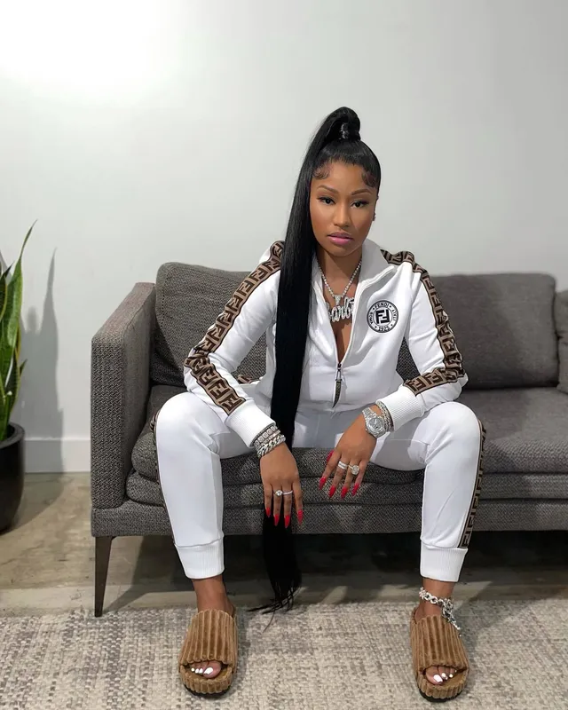 Nicki Minaj sitting on a sofa in a white tracksuit and metallic slides.