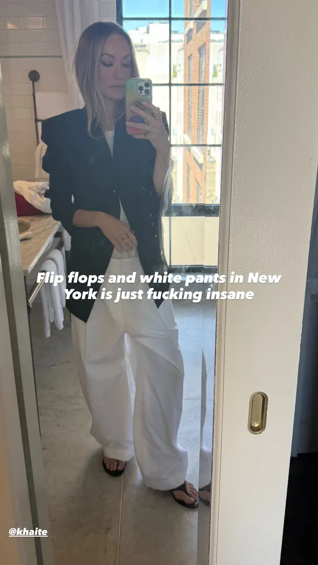Olivia Wilde posing for a mirror photograph in white trousers and black flip-flops.