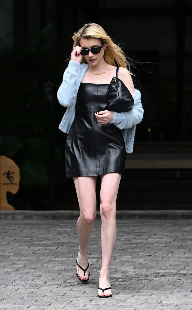 Emma Roberts walking outdoors in a black dress and flat sandals.