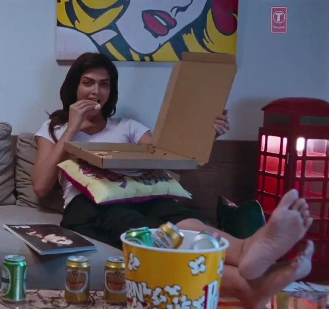 Deepika Padukone resting her bare feet on a table while holding a pizza box in a screen scene.