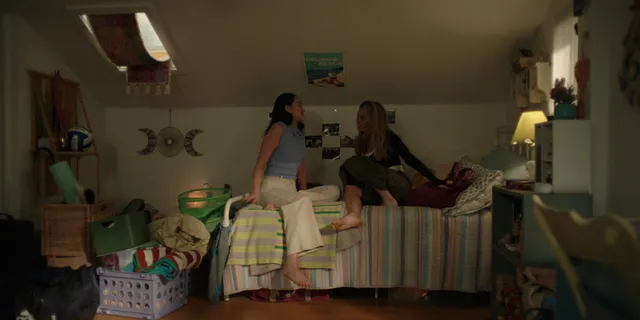 Lola Tung sitting on a bed in a blue top and pale skirt.
