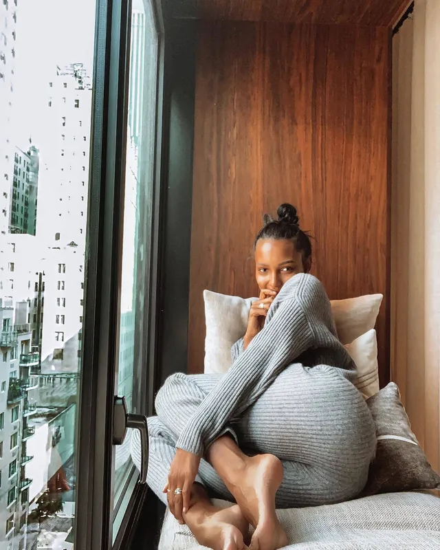 Jasmine Tookes sitting on a bed with her bare soles visible in a grey knit outfit.
