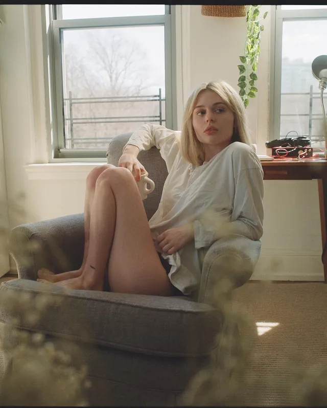 Emily Alyn Lind sitting barefoot in an armchair beside a window.