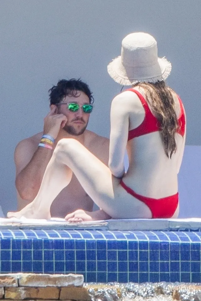 Hailee Steinfeld sitting barefoot beside a pool in a red swimsuit.