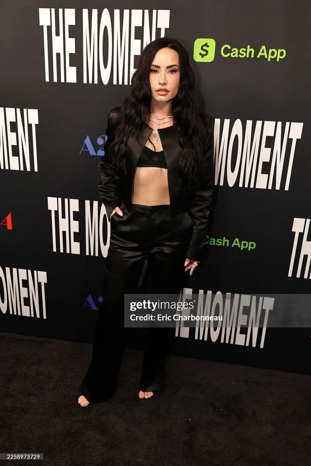 Demi Lovato standing at an event in a black outfit and open-toe footwear.