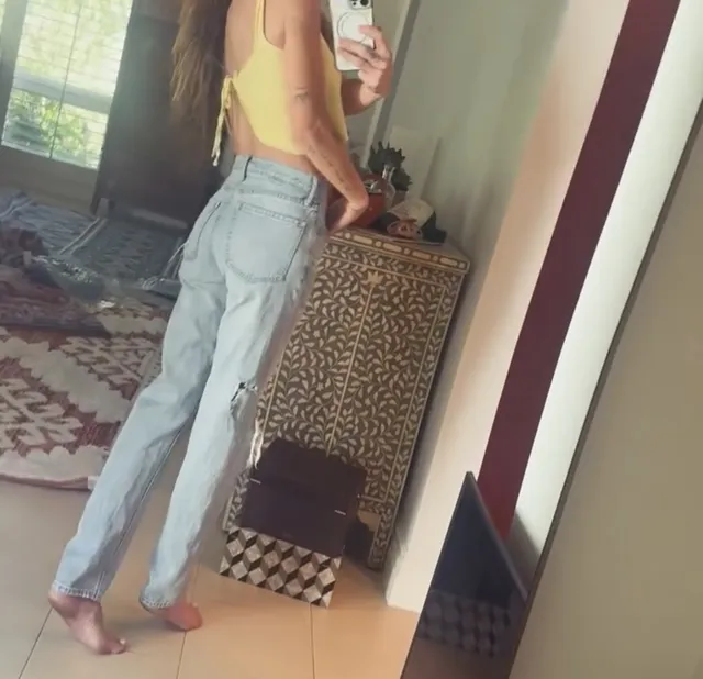 Sarah Shahi taking a mirror portrait in jeans and a light top.