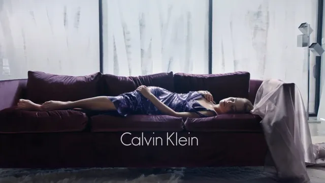 Margot Robbie reclining barefoot on a purple sofa in a fashion photograph.