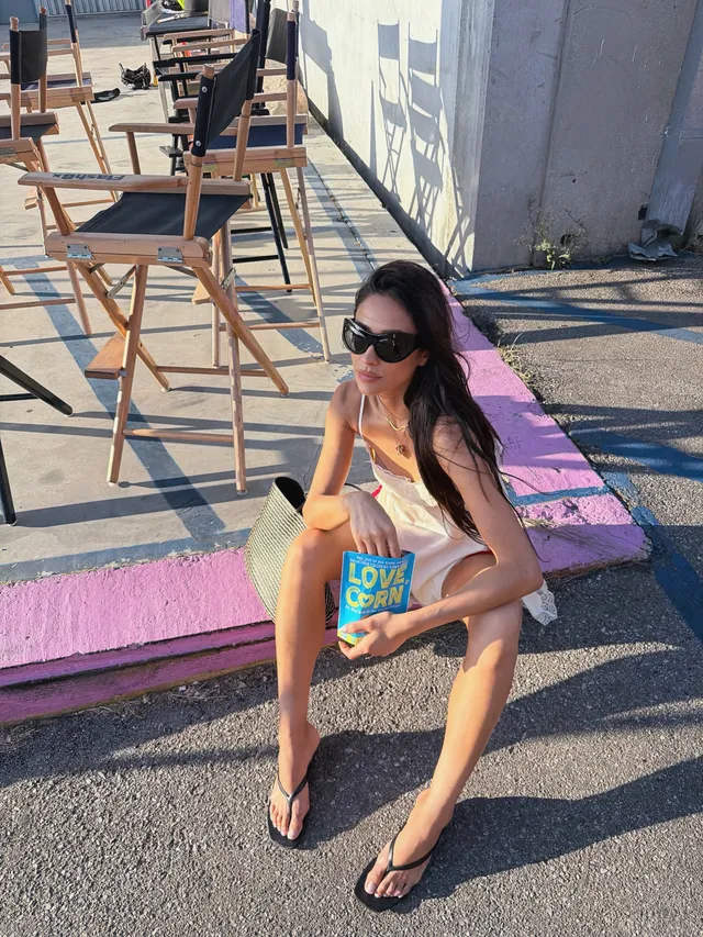 Shay Mitchell sitting on a street curb wearing flip-flops.