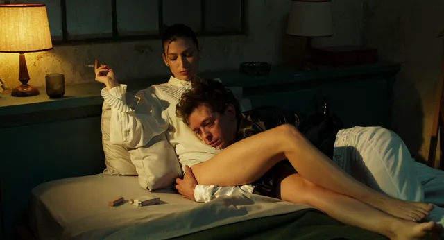 Úrsula Corberó reclining barefoot beside a man on a bed in a film scene.