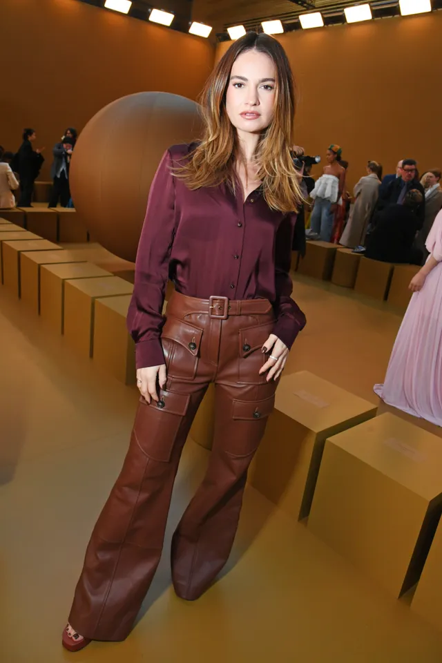 Lily James standing indoors in brown trousers and heels.