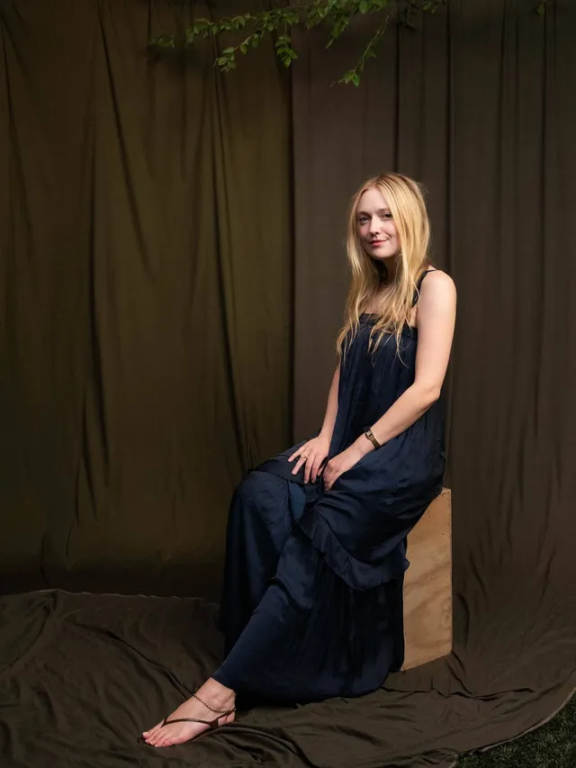 Dakota Fanning posing in a dark outfit and sandals against a brown backdrop.
