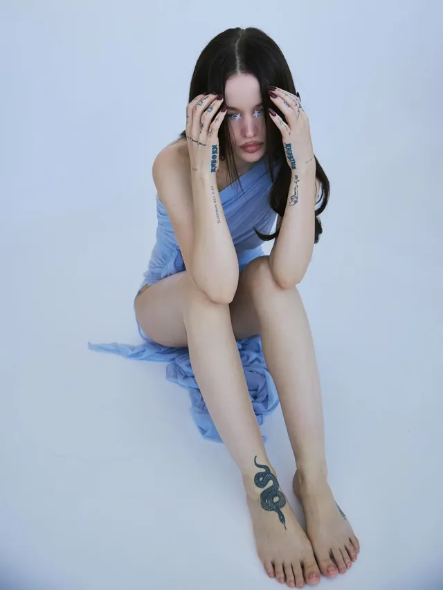 Dove Cameron sitting barefoot in a pale blue studio dress.