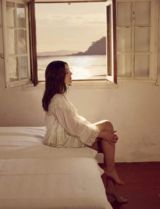 Monica Bellucci seated on a bed beside an open seaside window.
