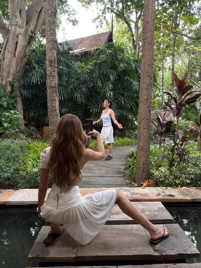 Lisa (Lalisa Manobal) sitting on a wooden terrace in dark flat sandals while a companion takes a photograph.