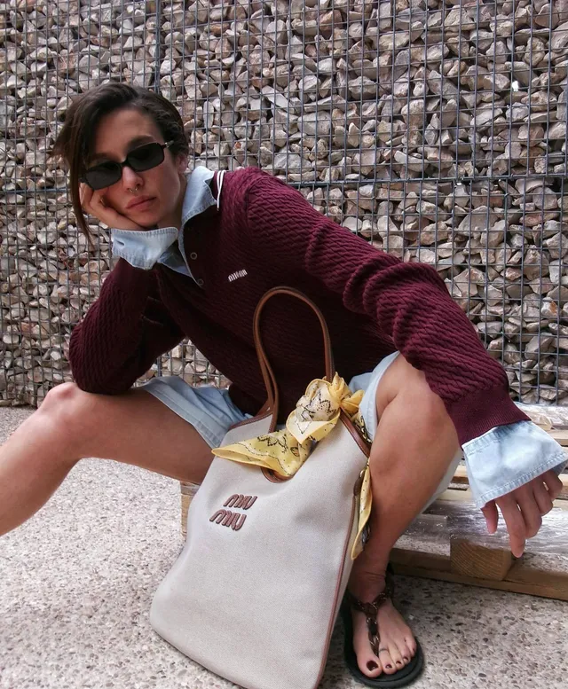 María Pedraza crouching outdoors in a burgundy sweater, shorts and dark flip-flops.