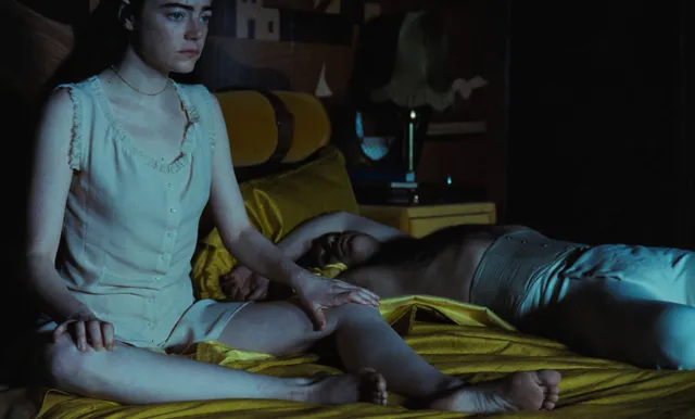 Emma Stone sitting on a bed with her bare legs and feet extended.