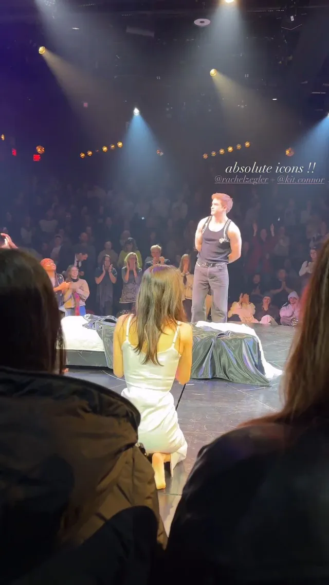 Rachel Zegler kneeling barefoot onstage among other performers.