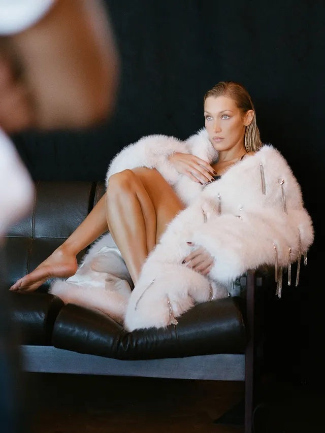 Bella Hadid sitting barefoot on a chair in a pale coat.