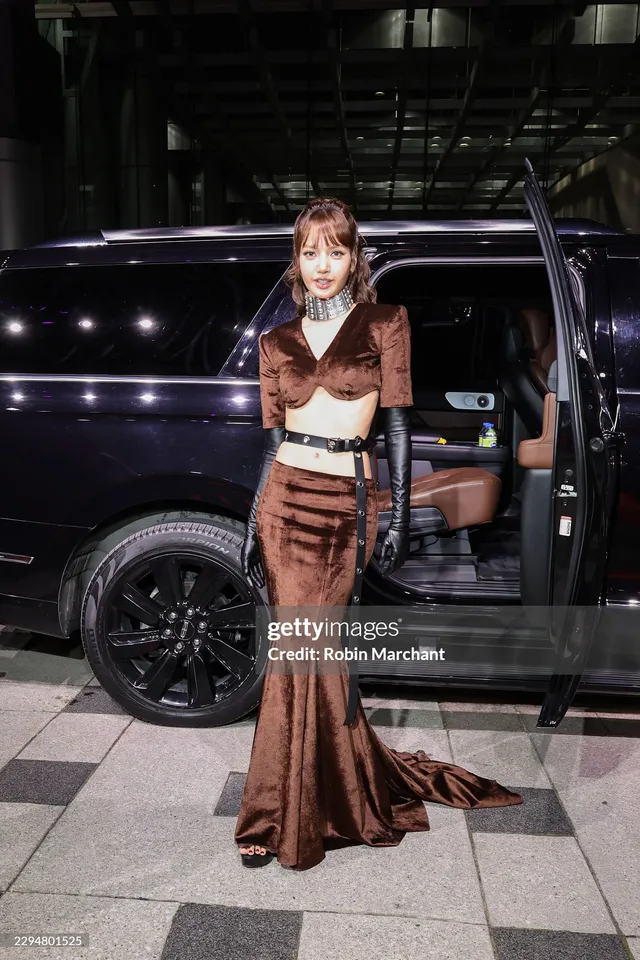 Lisa (Lalisa Manobal) wearing heels beneath a long brown outfit beside a vehicle.