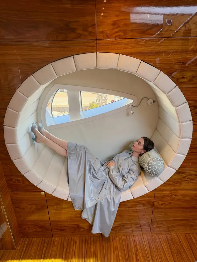 Lily Collins reclining in a circular window wearing a pale dress.