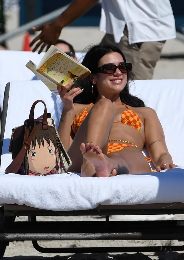 Dua Lipa: seated barefoot on a lounger holding a drink.