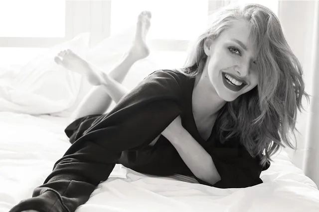 Amanda Seyfried lying barefoot on a white bed in a monochrome portrait.