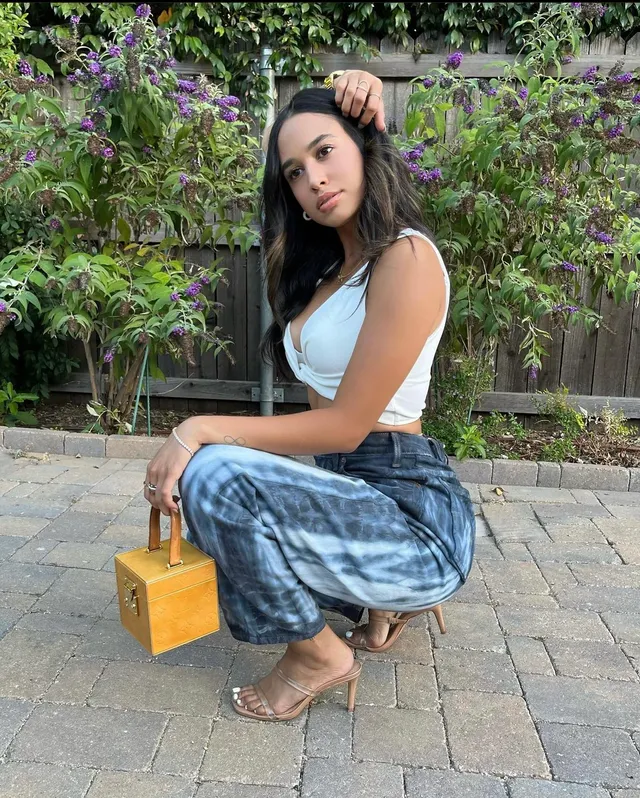 Emily Tosta crouching outdoors in jeans and clear heels.